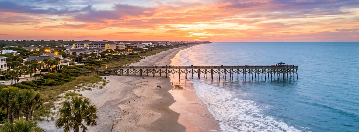 Aerial view of the Isle of Palms coastline at sunset featuring a long wooden fishing pier, beachfront luxury hotels, and the Atlantic Ocean waves in South Carolina.
